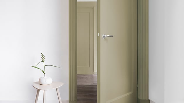 Een minimalistische gang met een lichtgroene open deur en een krukje met een witte vaas en een enkele plantstengel.