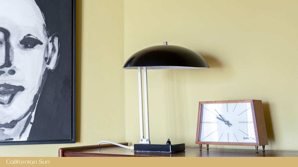Close‑up van een dressoir met een zwarte tafellamp, retro klok en een gedeeltelijk zichtbaar zwart‑wit schilderij tegen een gele muur