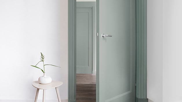 Een minimalistische gang met een open lichtgroene deur en een klein tafeltje met een witte vaas met een enkele plantstengel.