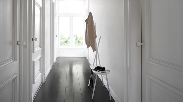 Een lichte, minimalistische gang met witte muren, donkere vloer en een krukje met een jas en accessoires.